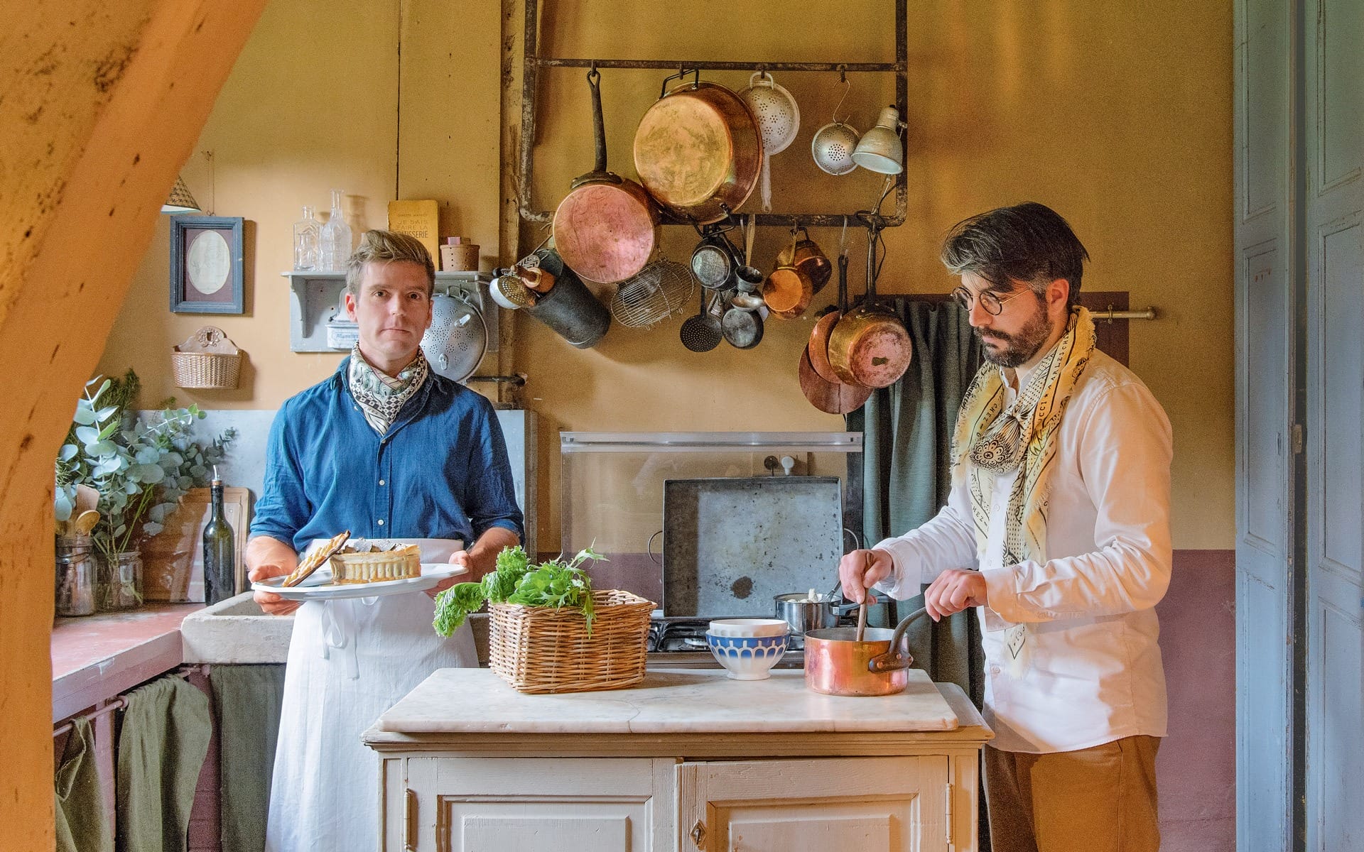 Two men cooking in a rustic kitchen, one holding a pie, the other stirring a pot, with hanging copper cookware.