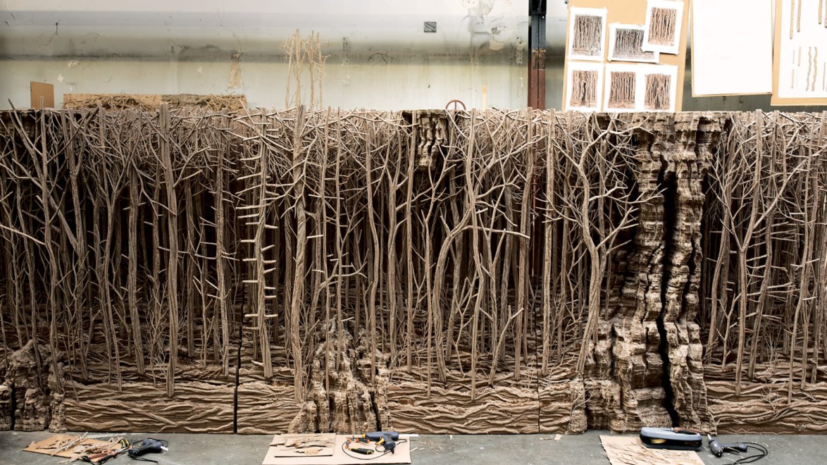 Intricate cardboard forest art installation in a studio with tools and sketches visible in the background.