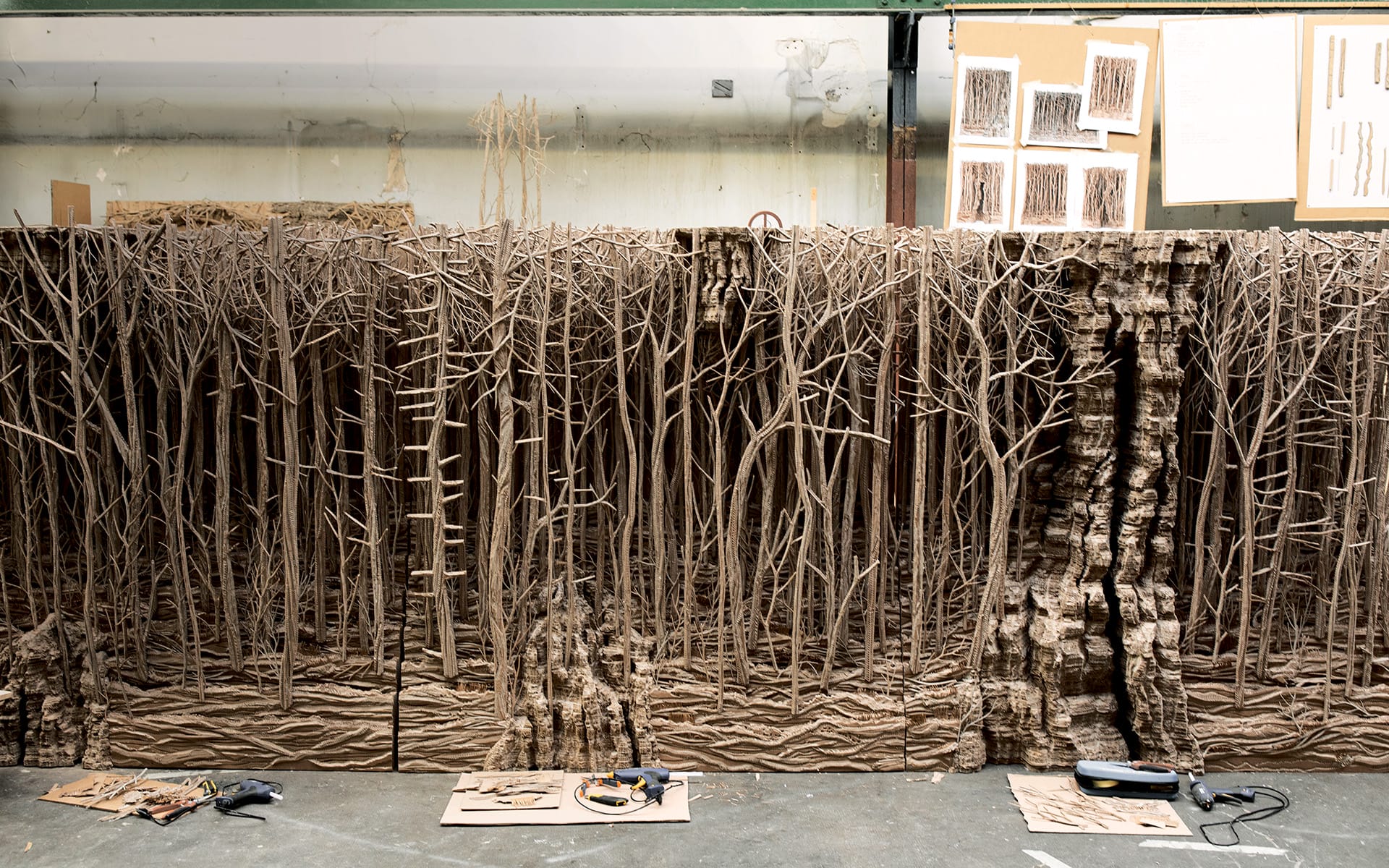 Intricate cardboard forest art installation in a studio with tools and sketches visible in the background.