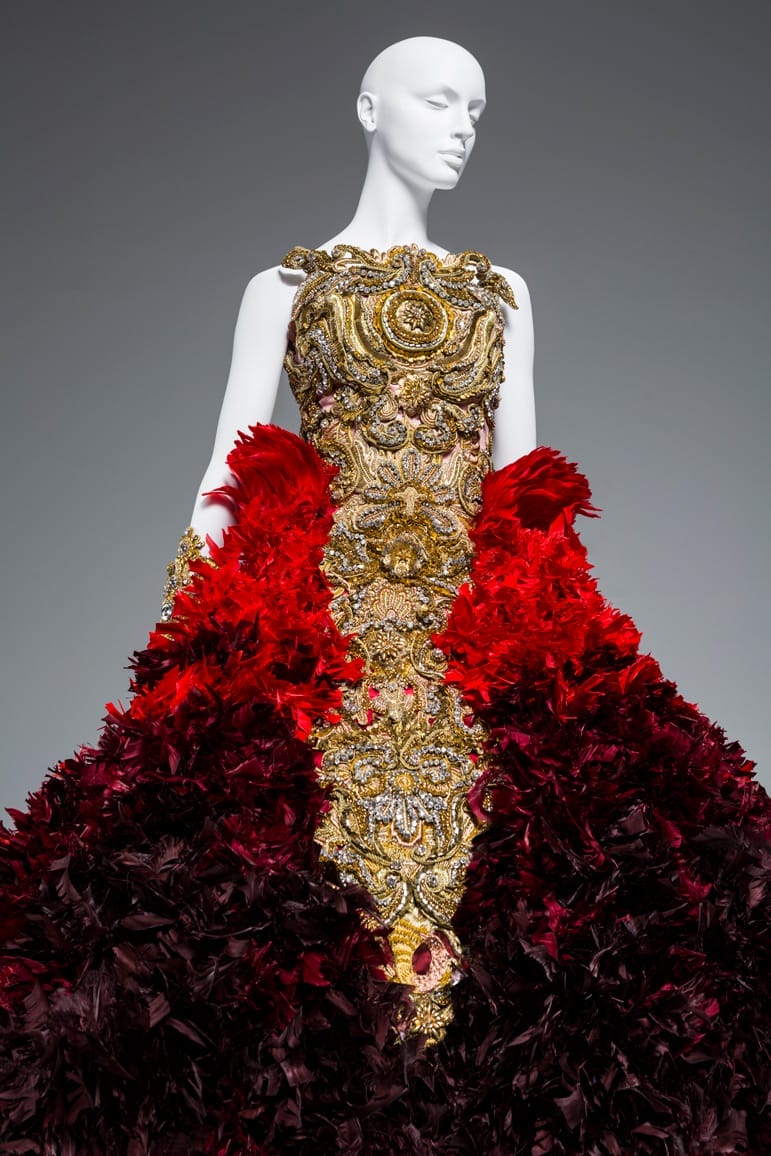 Mannequin wearing an intricate gold and red feather gown with ornate detailing on a neutral background.