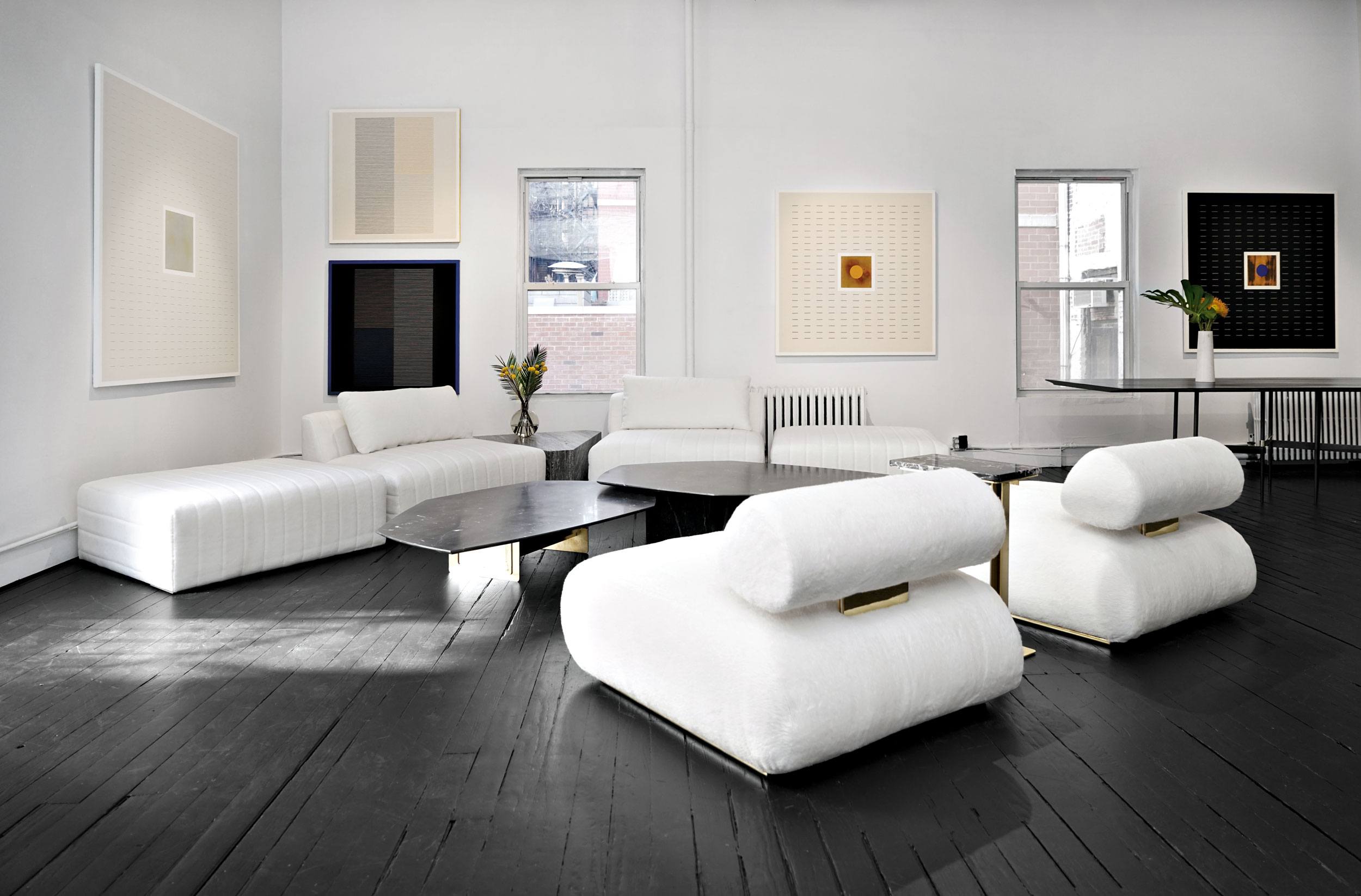 Modern art gallery interior with white couches, abstract wall art, dark wood flooring, and large windows.