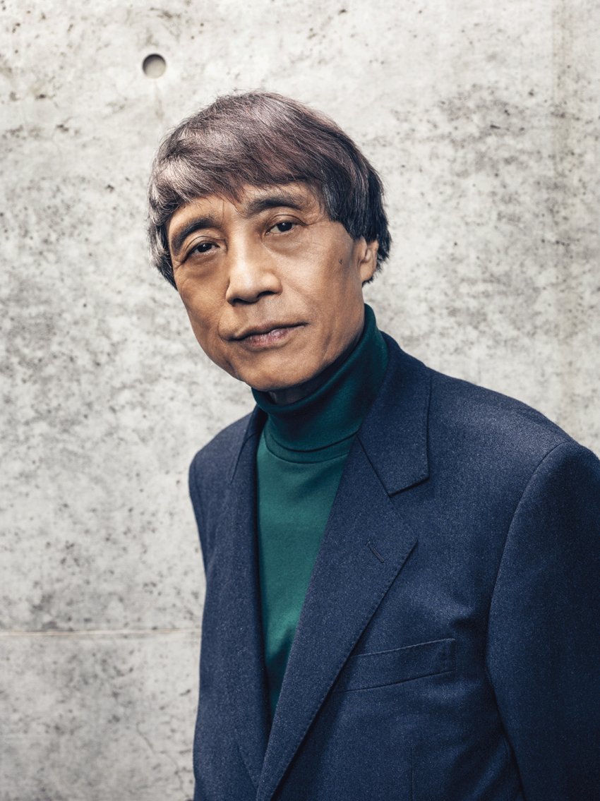 Person in a blue suit and green turtleneck standing against a concrete wall