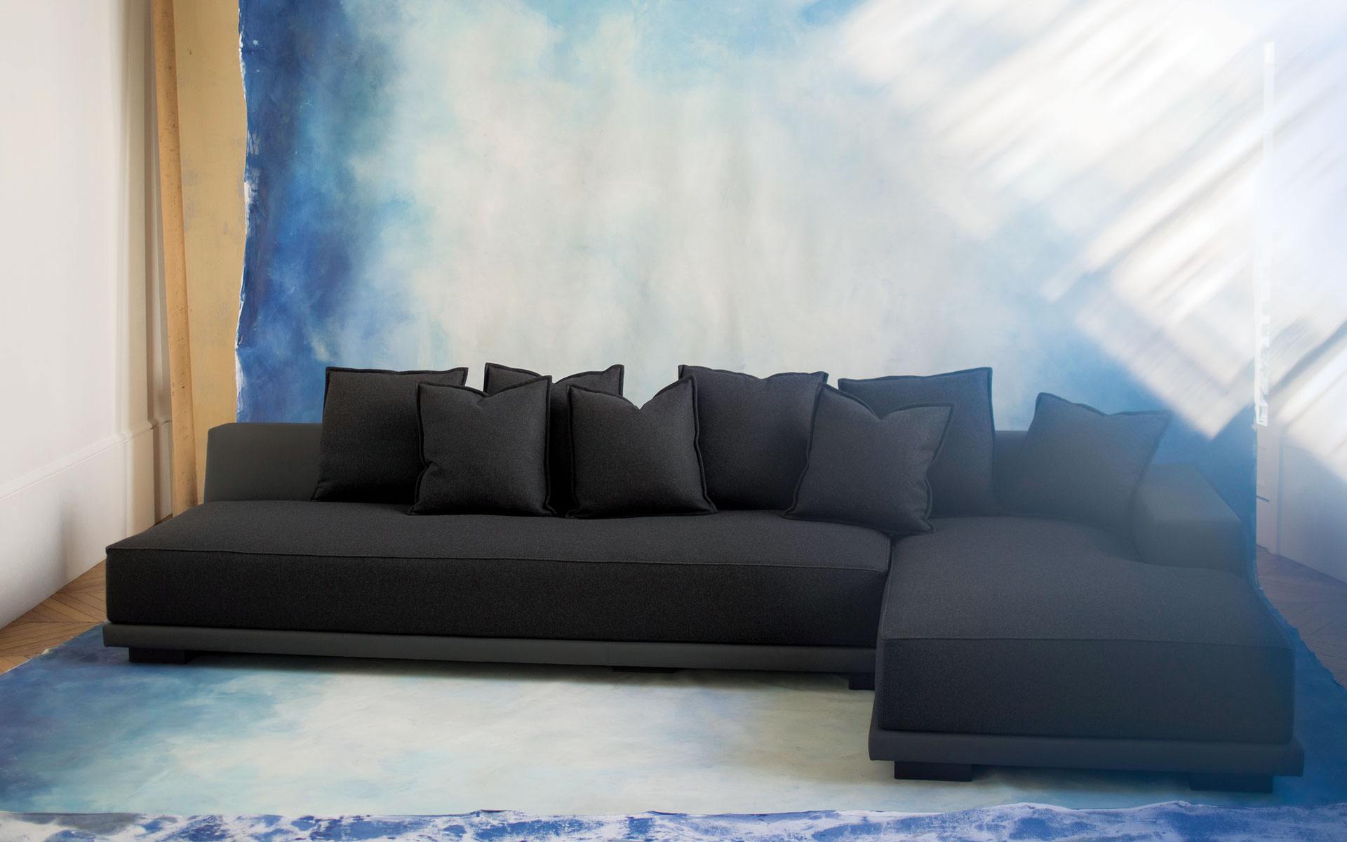 Black sectional sofa with multiple cushions against a blue abstract wall in a sunlit room.