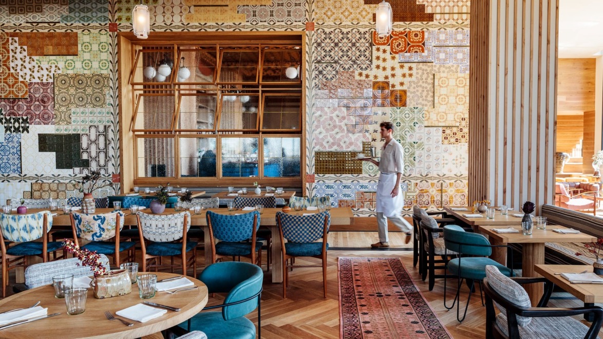 Colorful restaurant interior with patterned walls, wooden tables, and chairs; a waiter walking through the dining area.