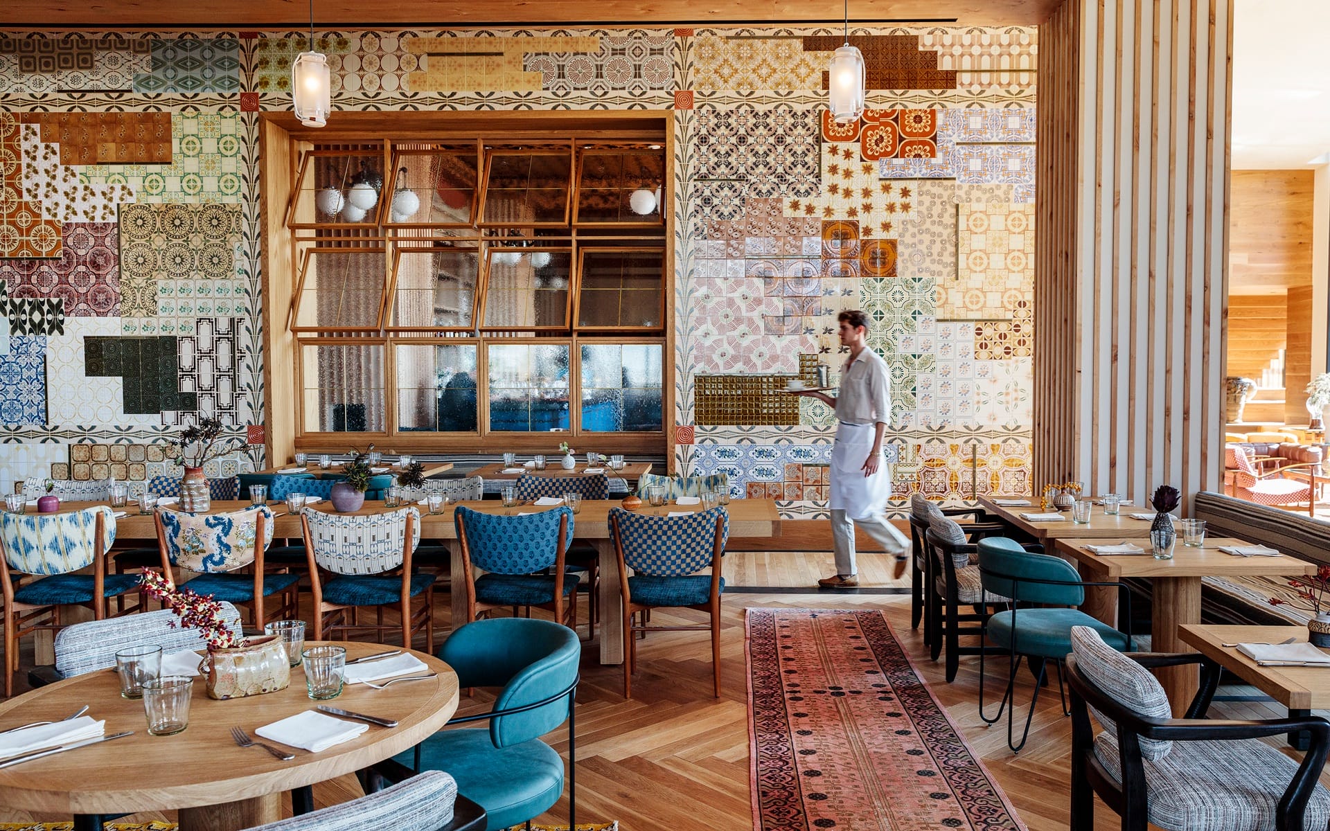Colorful restaurant interior with patterned walls, wooden tables, and chairs; a waiter walking through the dining area.