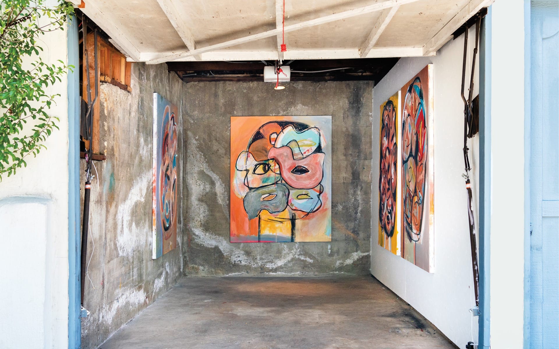 Colorful abstract paintings displayed in a small, rustic garage with concrete walls and natural light streaming in.