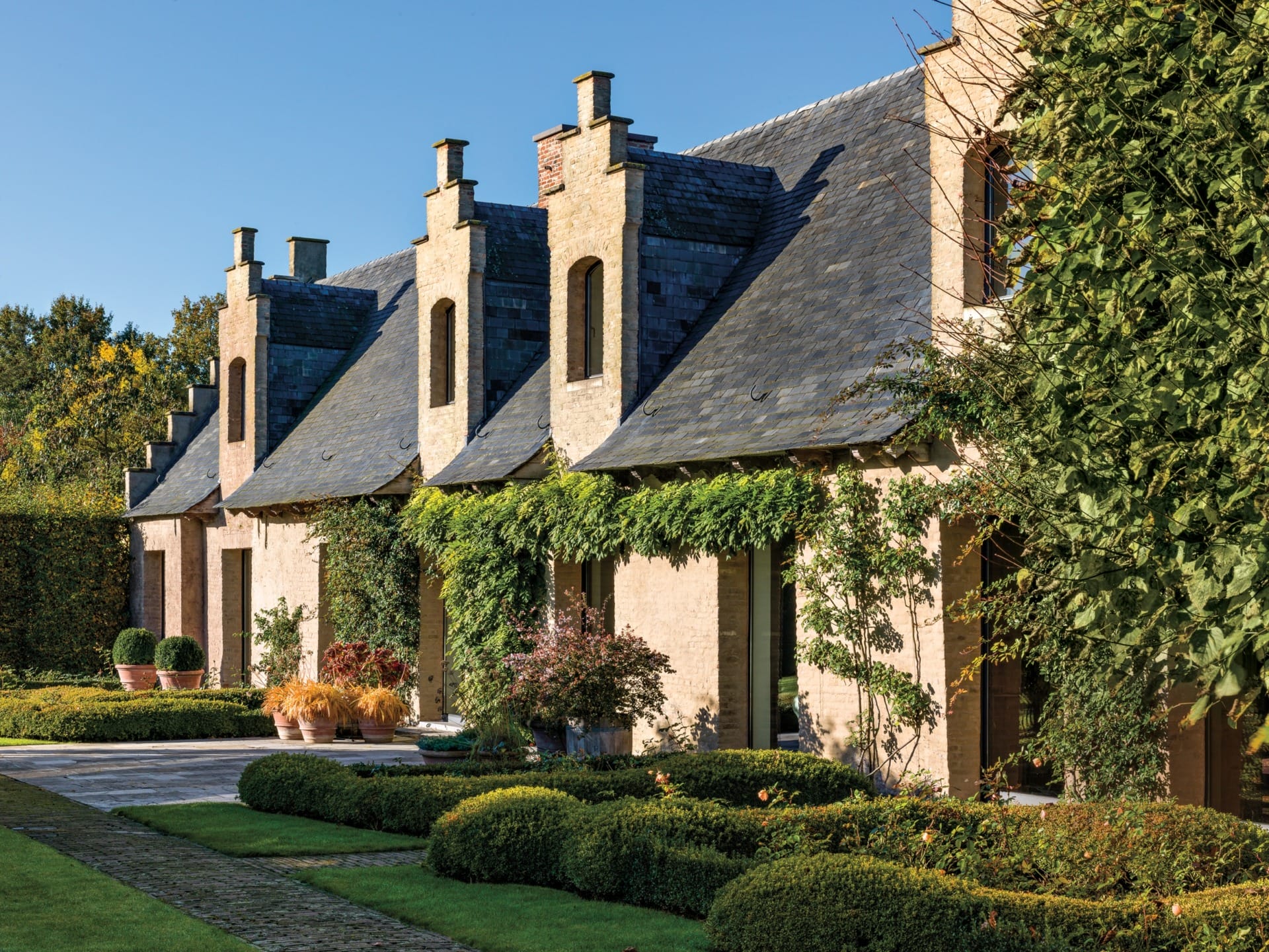 Charming house with ivy-covered brick exterior, slate roof, and trimmed hedges in a sunny landscaped garden setting.