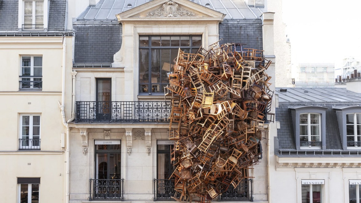 Art installation of numerous chairs seemingly suspended in mid-air against the facade of a classic European-style building.