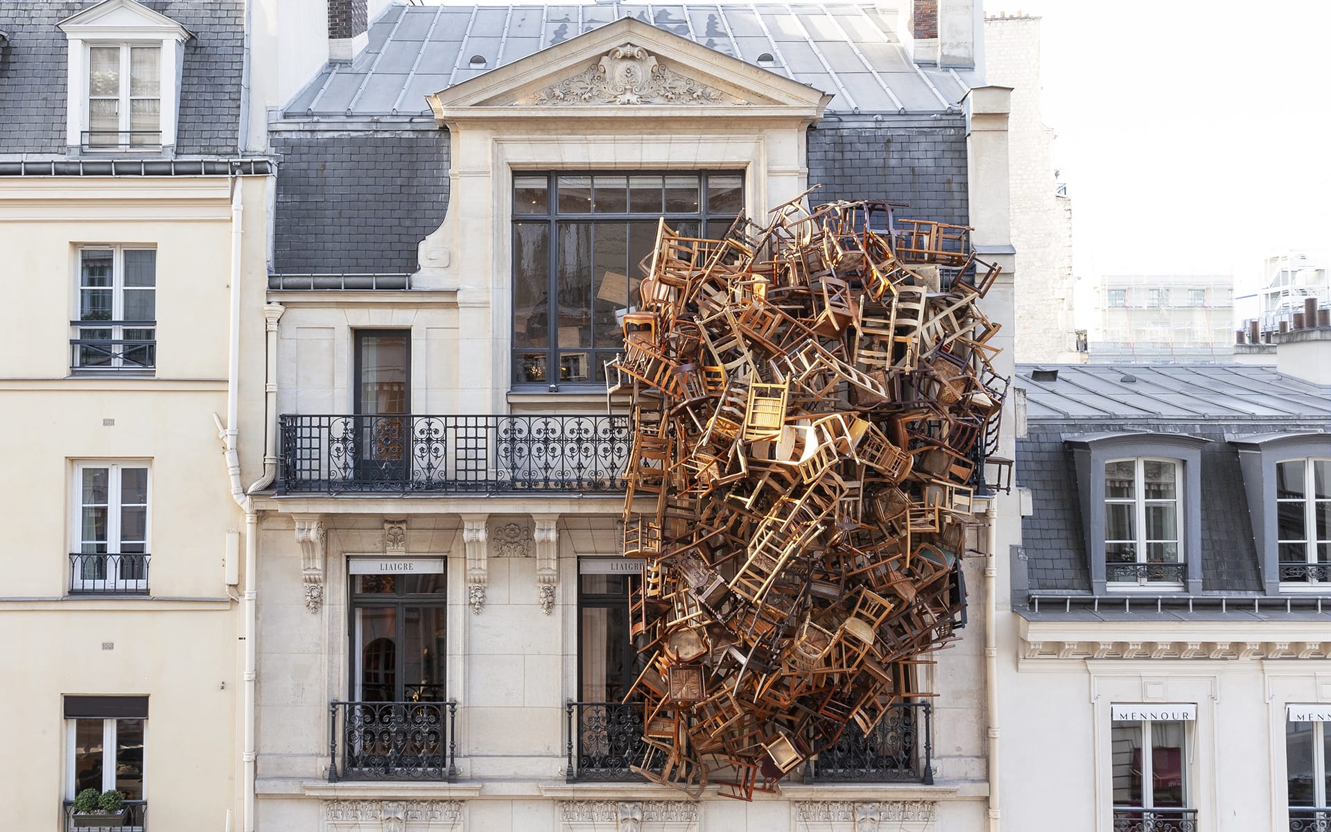 Art installation of numerous chairs seemingly suspended in mid-air against the facade of a classic European-style building.