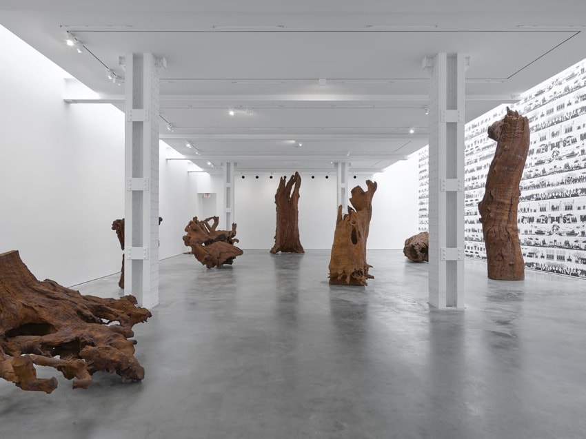 Contemporary art gallery with large wooden sculptures on a polished concrete floor and a black-and-white photo mural on the wall.