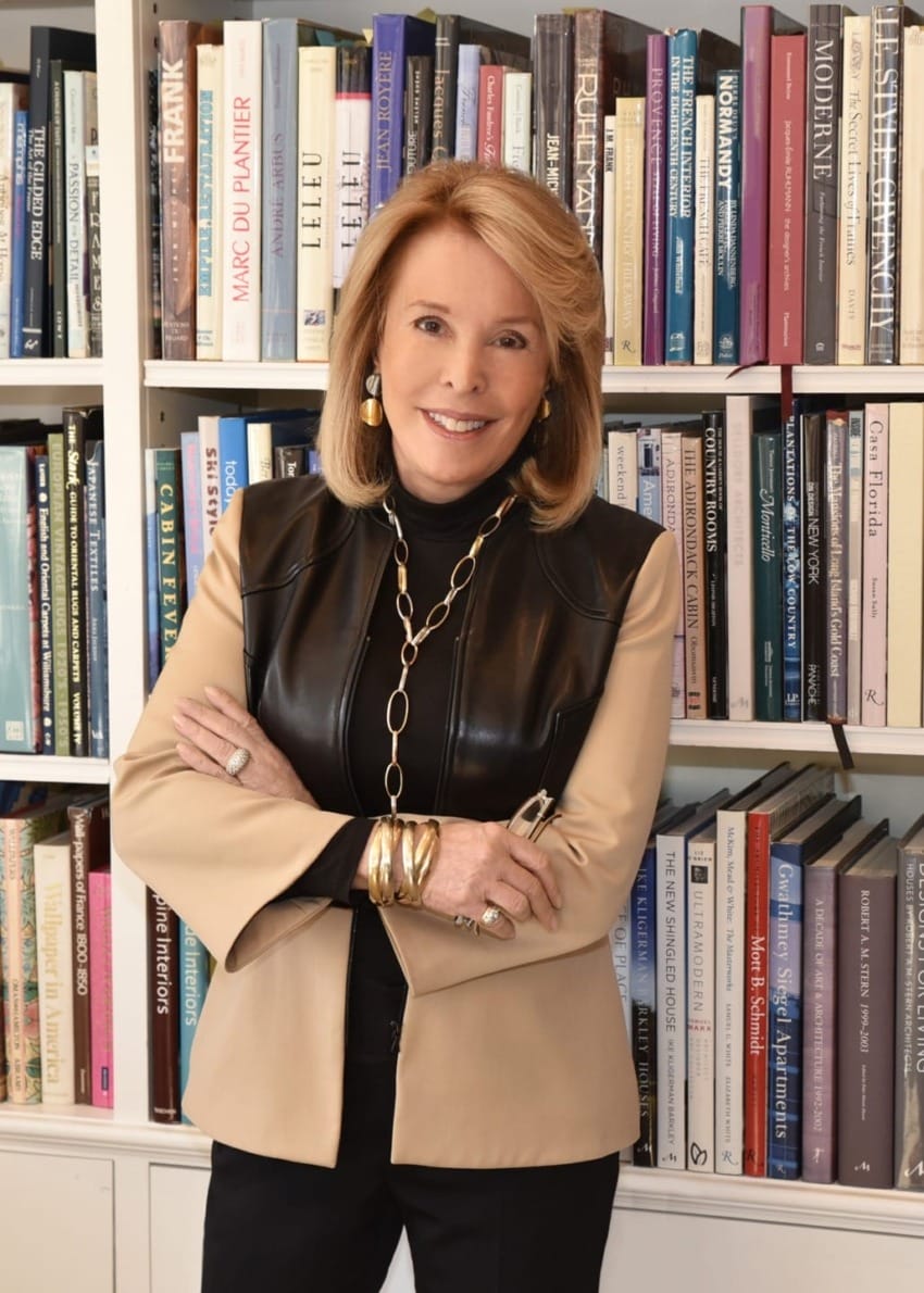 Person standing with folded arms in front of a bookshelf filled with various books.