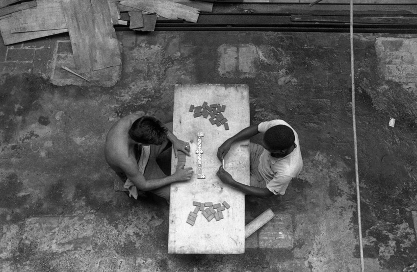 Two people playing a game with tiles on a table, view from above, surrounded by wooden boards and rustic ground.