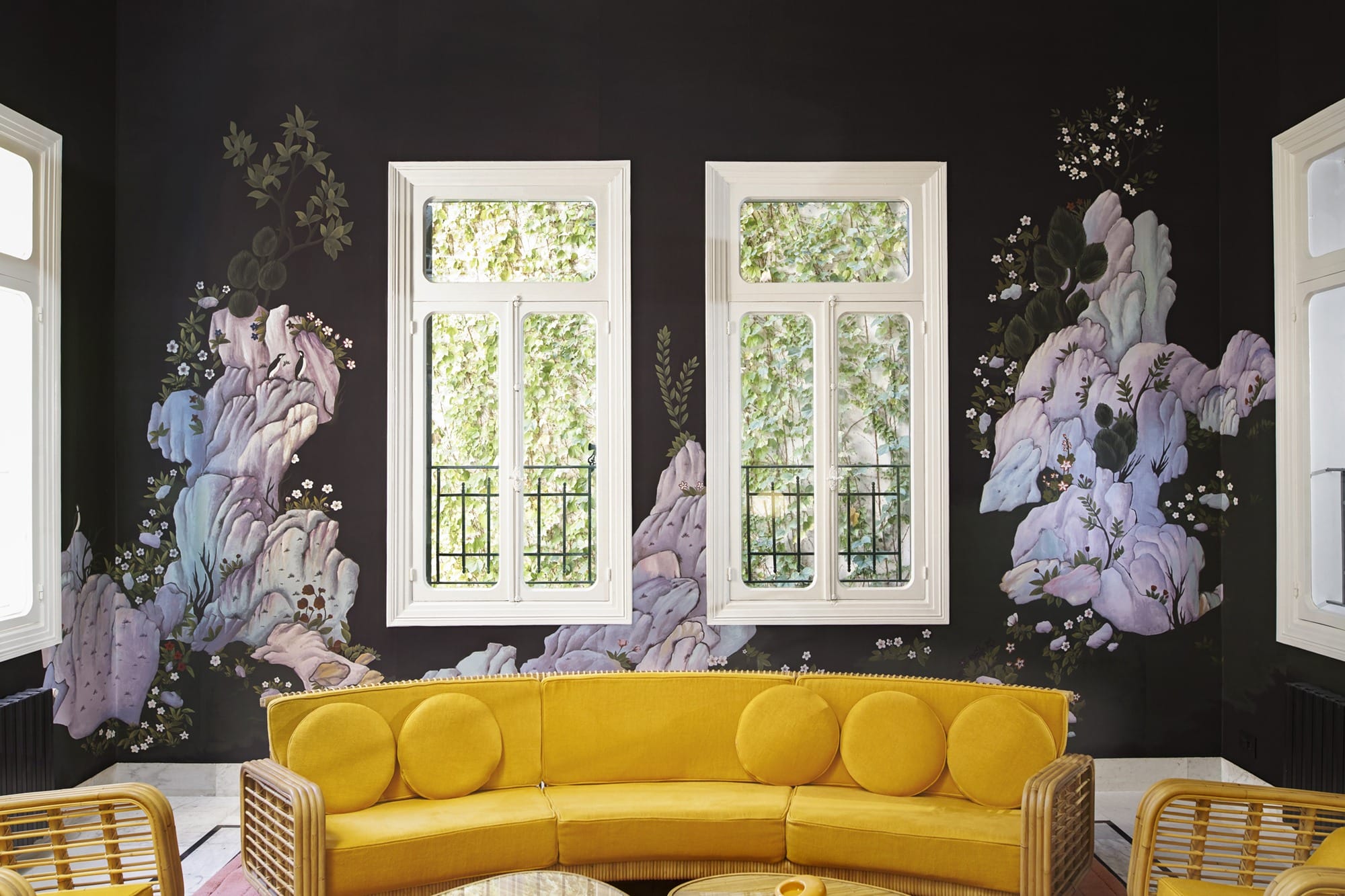 Yellow couch in front of a dark floral mural with two large windows, creating a vibrant and artistic interior space.