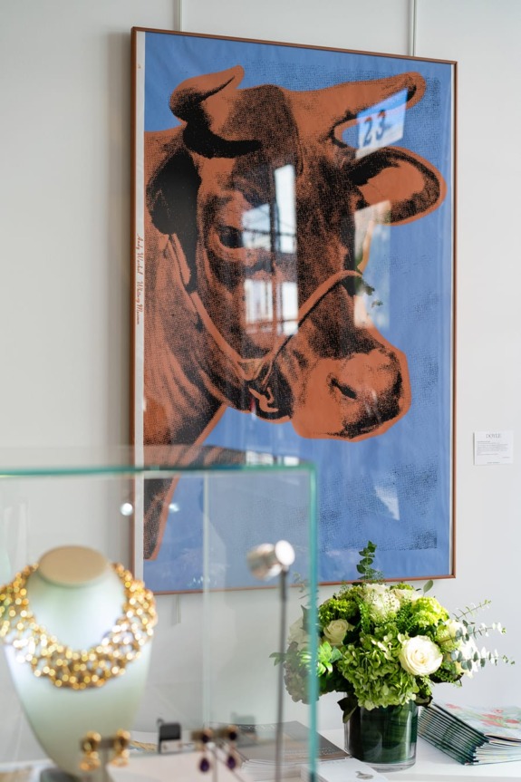 Cow-themed artwork on a wall, displayed above a glass case with jewelry and a vase of flowers.