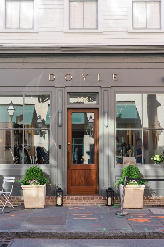 Front view of a stylish storefront with wooden door, large windows, and flower pots on the sidewalk.