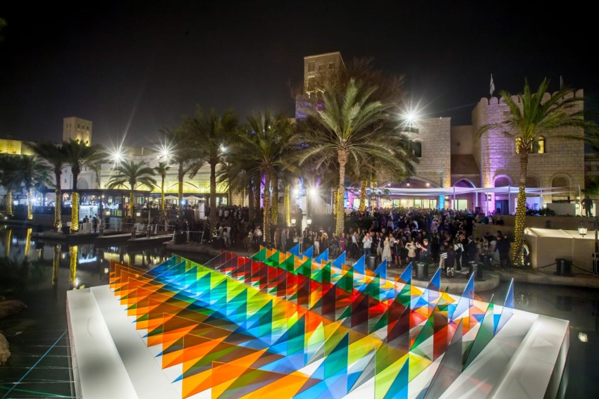 Nighttime view of a vibrant outdoor installation with colorful geometric patterns, many palm trees, and a crowd gathered.