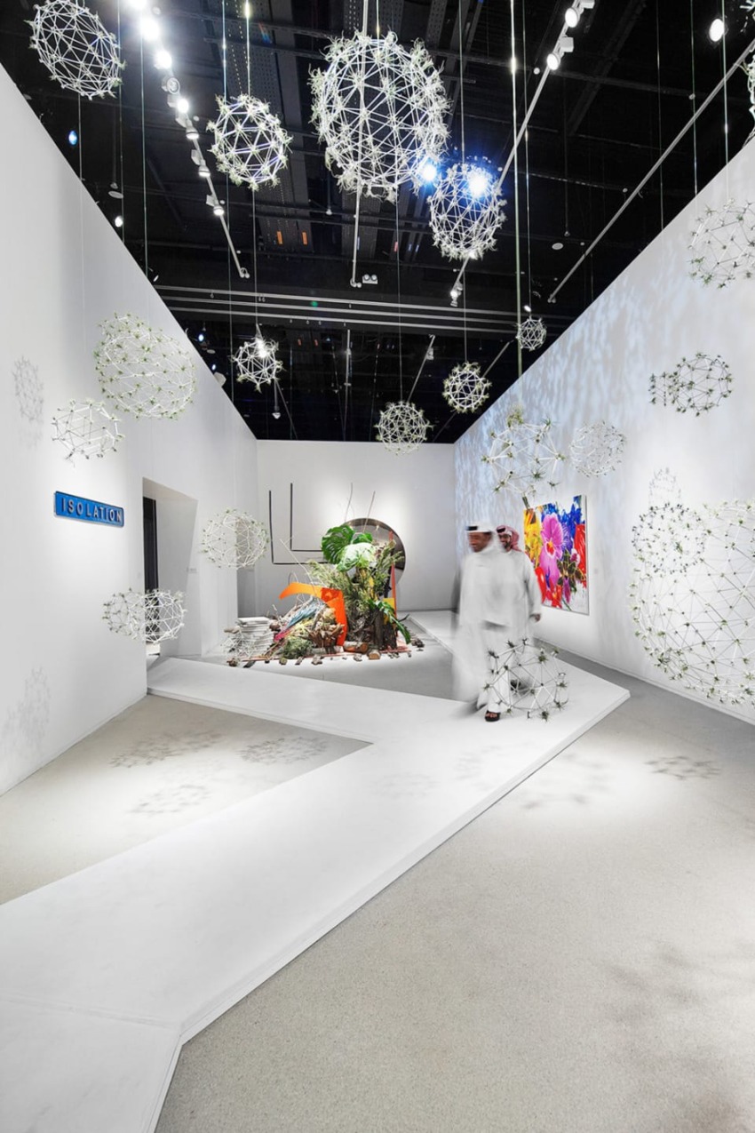 Art exhibit with hanging spherical sculptures, illuminated walkway, colorful abstract painting, and two people walking through.