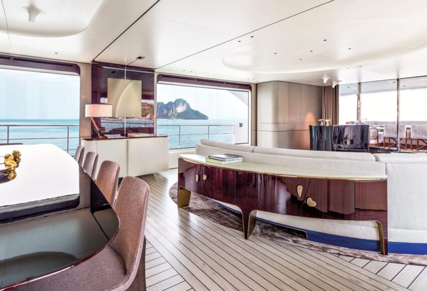 Luxury yacht interior with modern seating, dining table, and large windows showcasing ocean and island views.