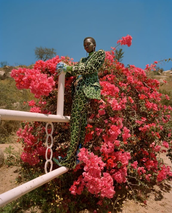 Person in a vibrant green leopard print suit stands amid bright pink flowers under a clear blue sky.