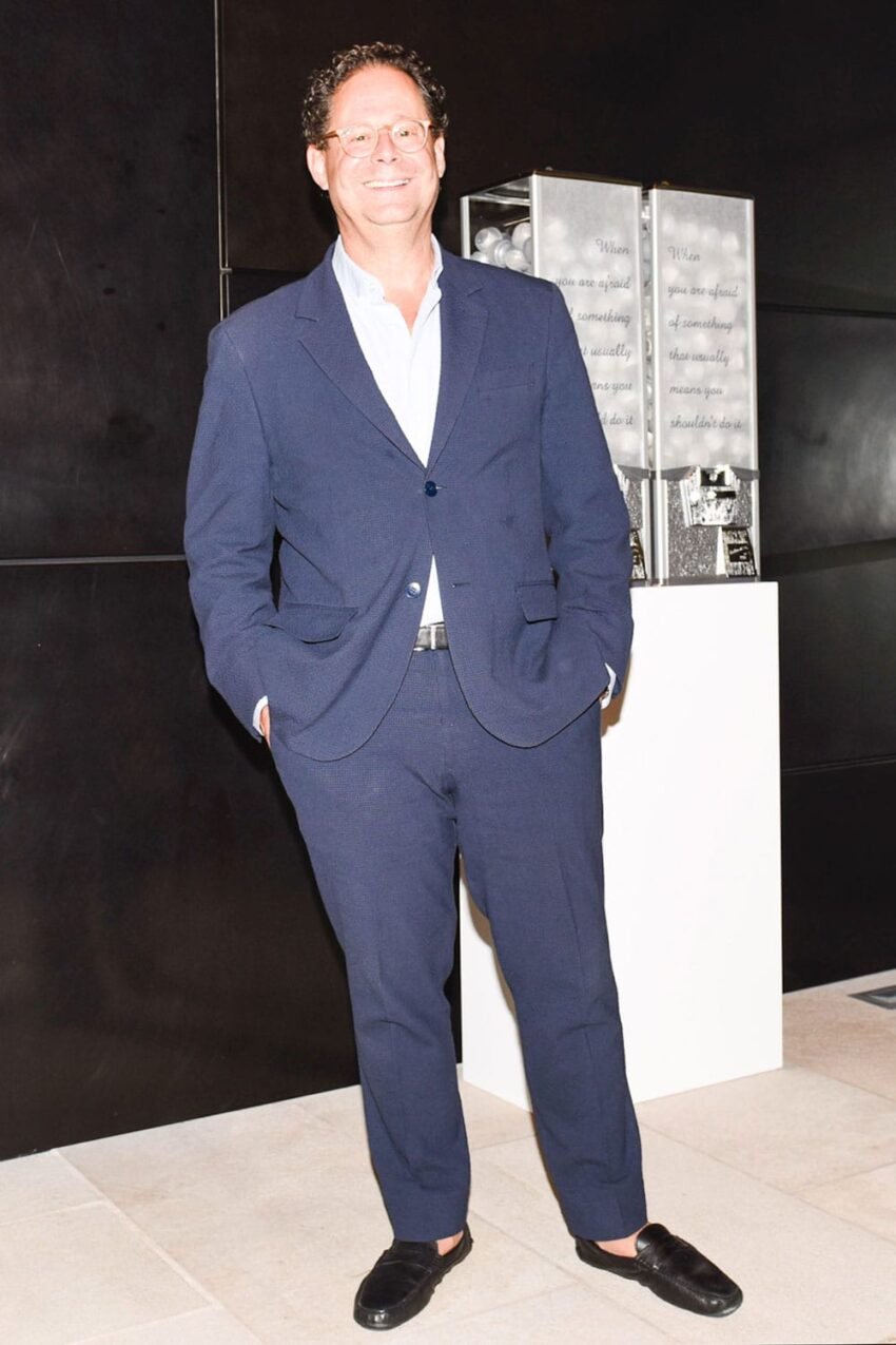 Man in a blue suit standing indoors with hands in pockets, smiling, two metal structures in the background.