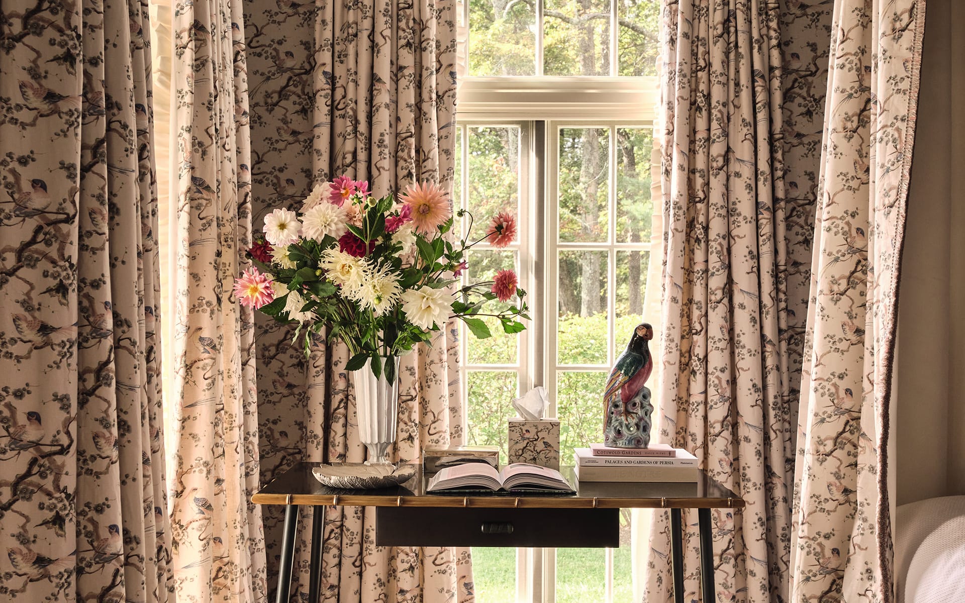 Vase of colorful flowers on table near window with floral curtains, open book, and decorative bird statue.