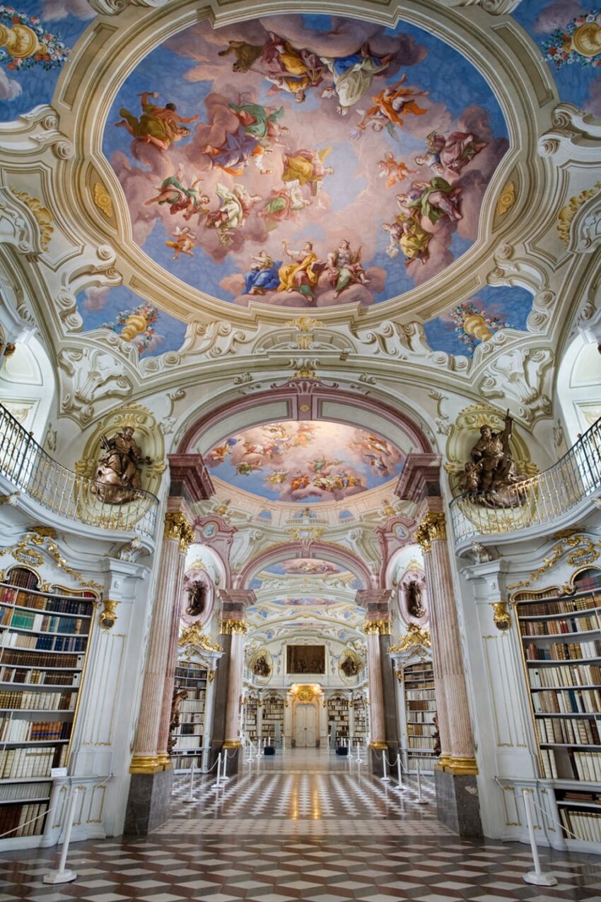 Ornate library interior with colorful ceiling fresco, tall bookshelves, and intricate architectural details.