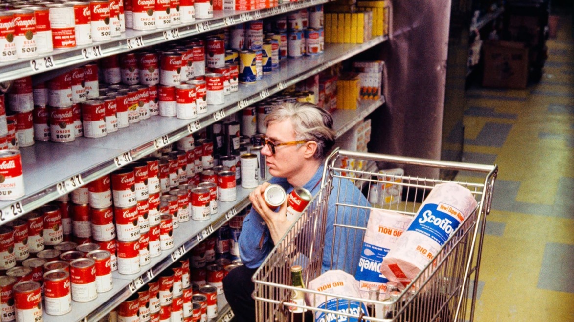 A person in a blue shirt browsing soup cans in a grocery store aisle with a shopping cart containing paper towels.