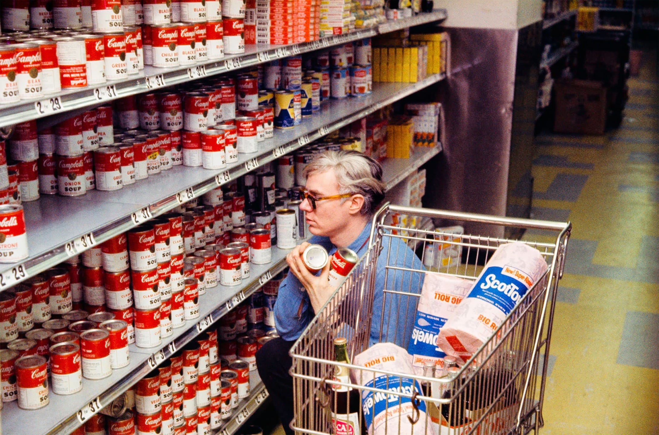 A person in a blue shirt browsing soup cans in a grocery store aisle with a shopping cart containing paper towels.