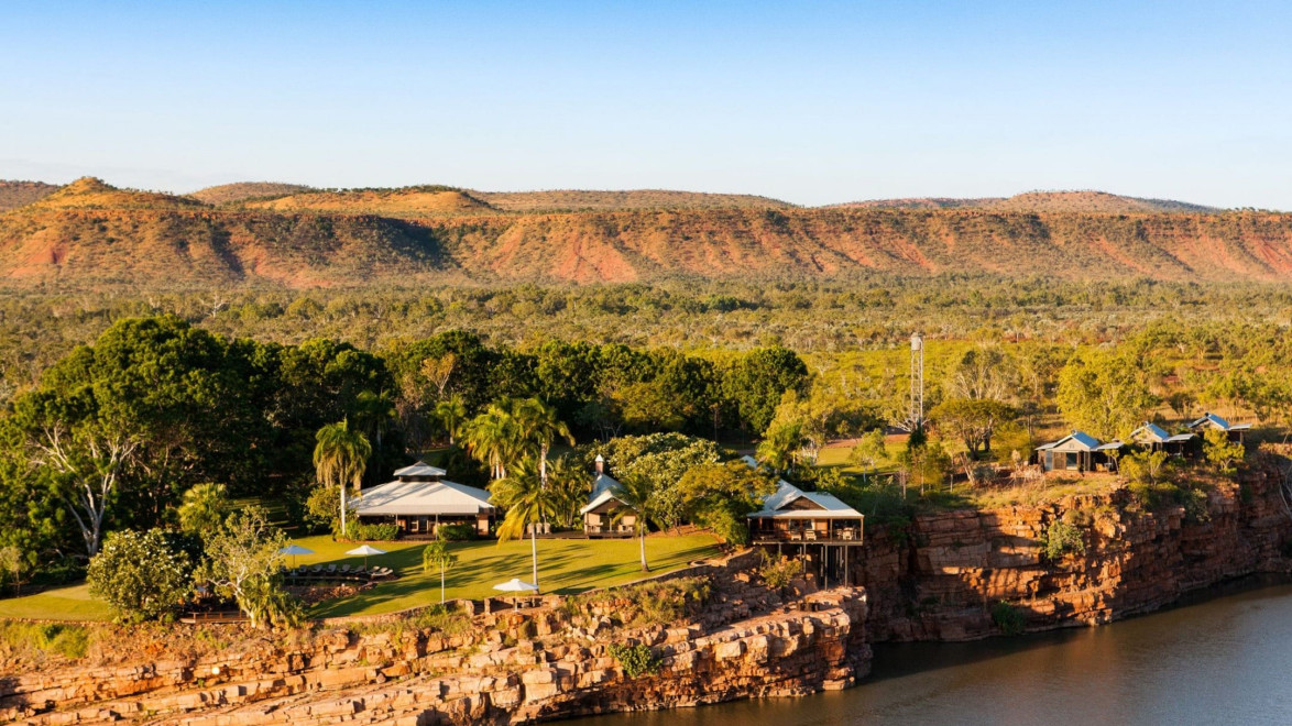 Aerial view of a remote resort on a rocky cliff with lush greenery, overlooking a calm river and distant hills under a clear sky.