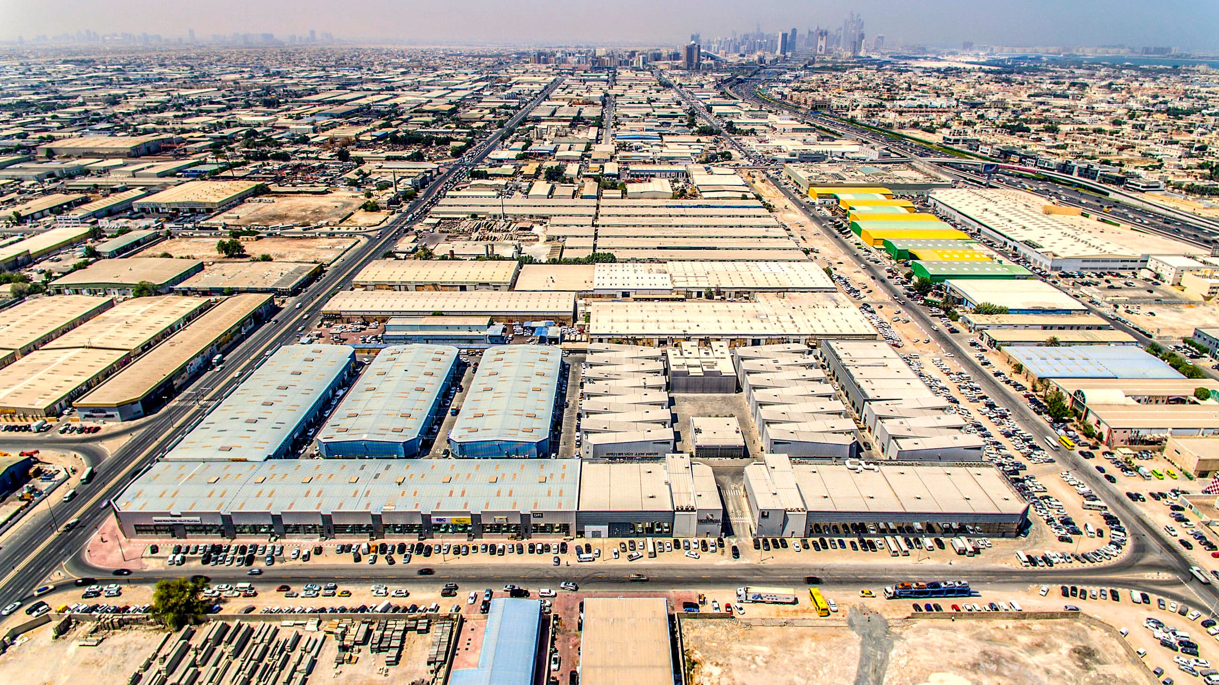 Aerial view of an extensive industrial area with warehouses and roads stretching into the distance, city skyline in background.