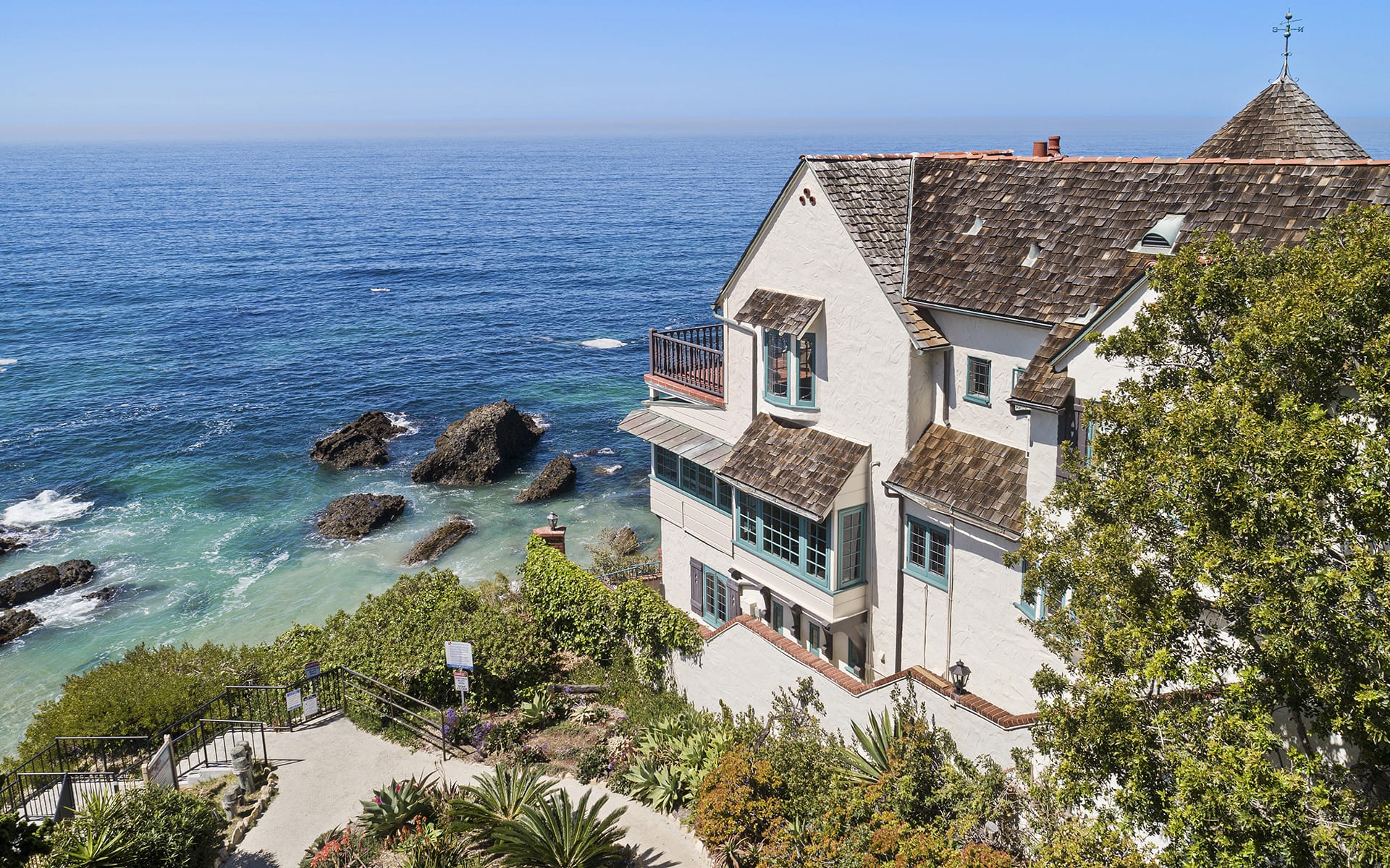 Clifftop house overlooking ocean with waves, surrounded by lush greenery and rocky shore.