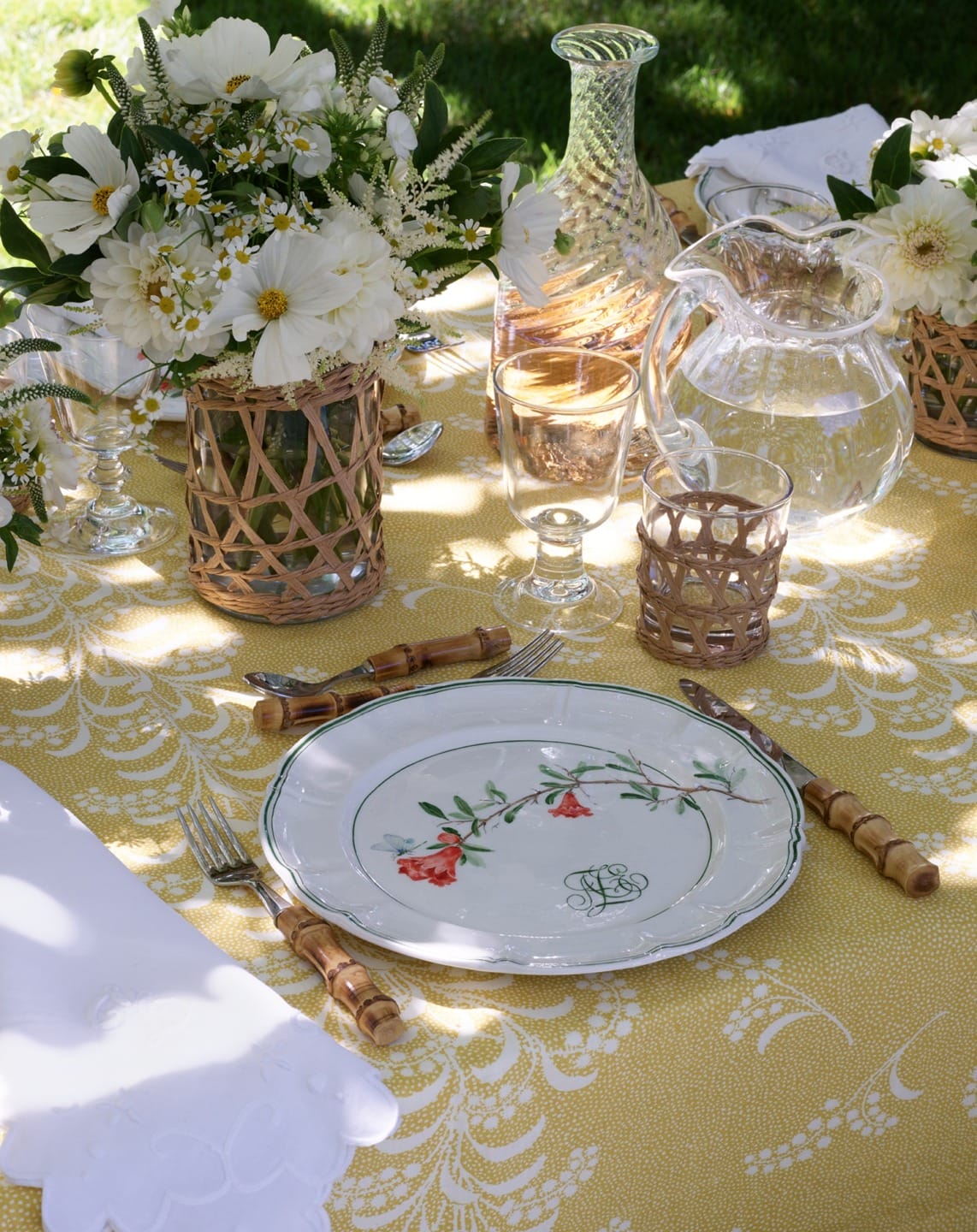 Elegant outdoor dining setup with floral arrangements, glassware, and a decorative plate on a yellow patterned tablecloth.