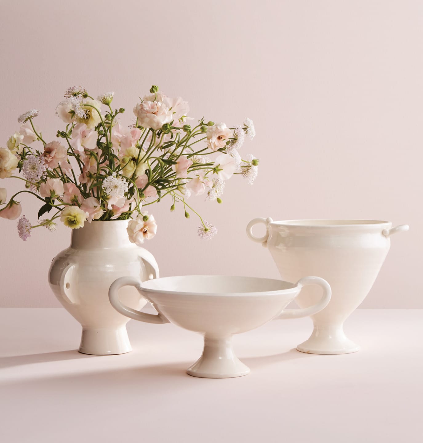 Three elegant white ceramic vases, one filled with delicate pink flowers, displayed on a soft pink background.