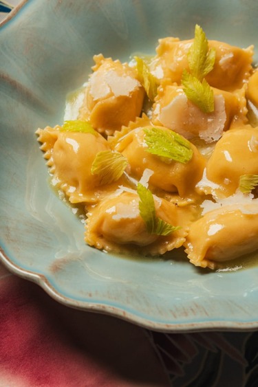 Plate of golden ravioli topped with parmesan and garnished with fresh celery leaves on a textured blue dish.