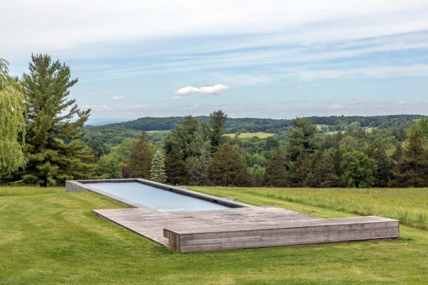 Rectangular outdoor pool surrounded by a wooden deck in a grassy landscape with trees and a cloudy sky.