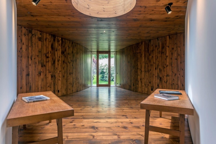 Wood-paneled hallway with wide planks and two wooden tables leading to glass door revealing outdoor greenery.