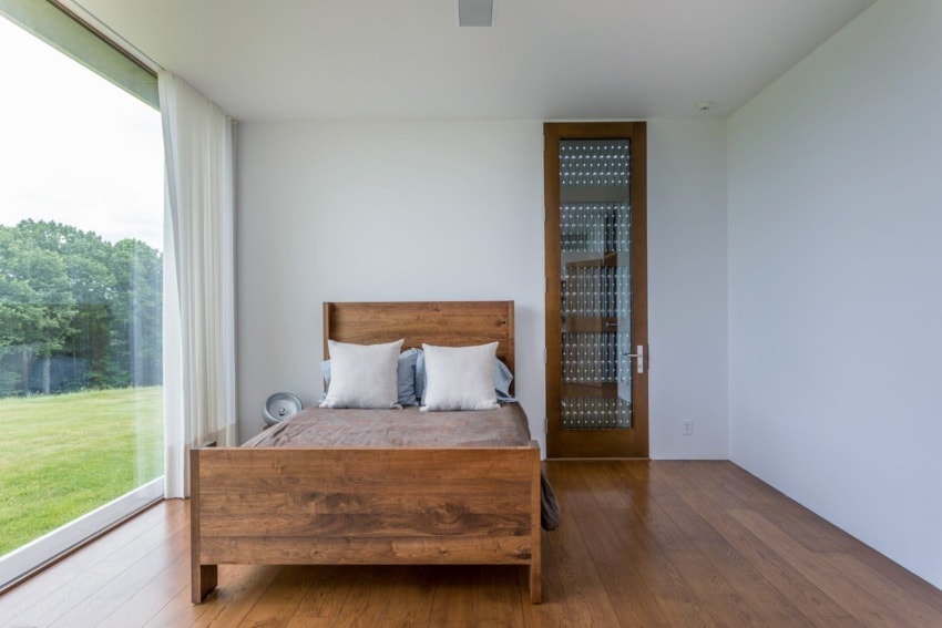 Minimalist bedroom with wooden bed frame, large window, and view of greenery outside.