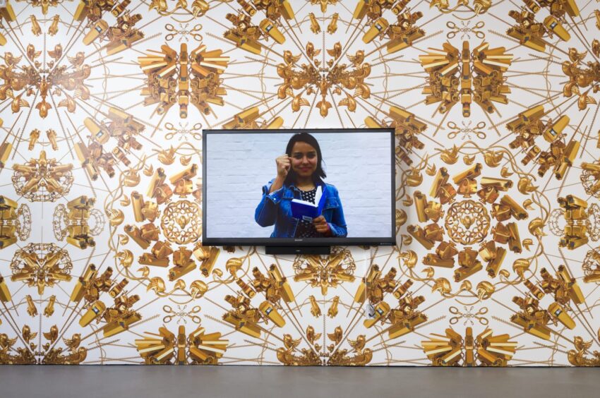 Television displaying a person on a patterned vintage wallpapered wall in a gallery setting.