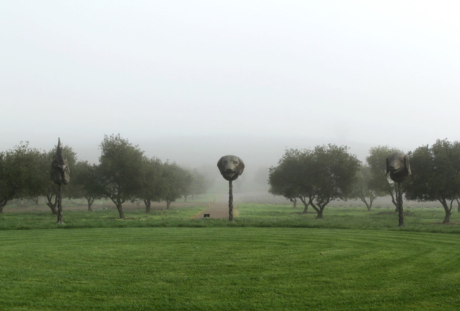 Three large horse head sculptures in a misty park landscape with green grass and surrounding trees.