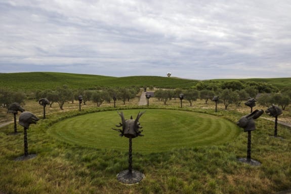 Sculptures of animal heads arranged in a circle on a grassy field with trees and hills in the background under a cloudy sky.
