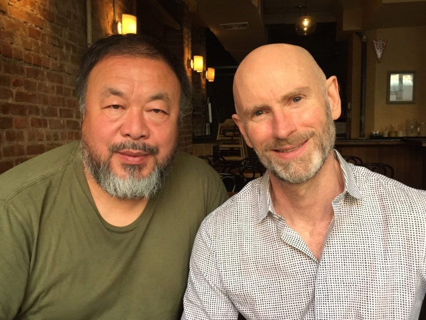 Two men sitting together in a dimly lit cafe with brick walls, smiling at the camera.