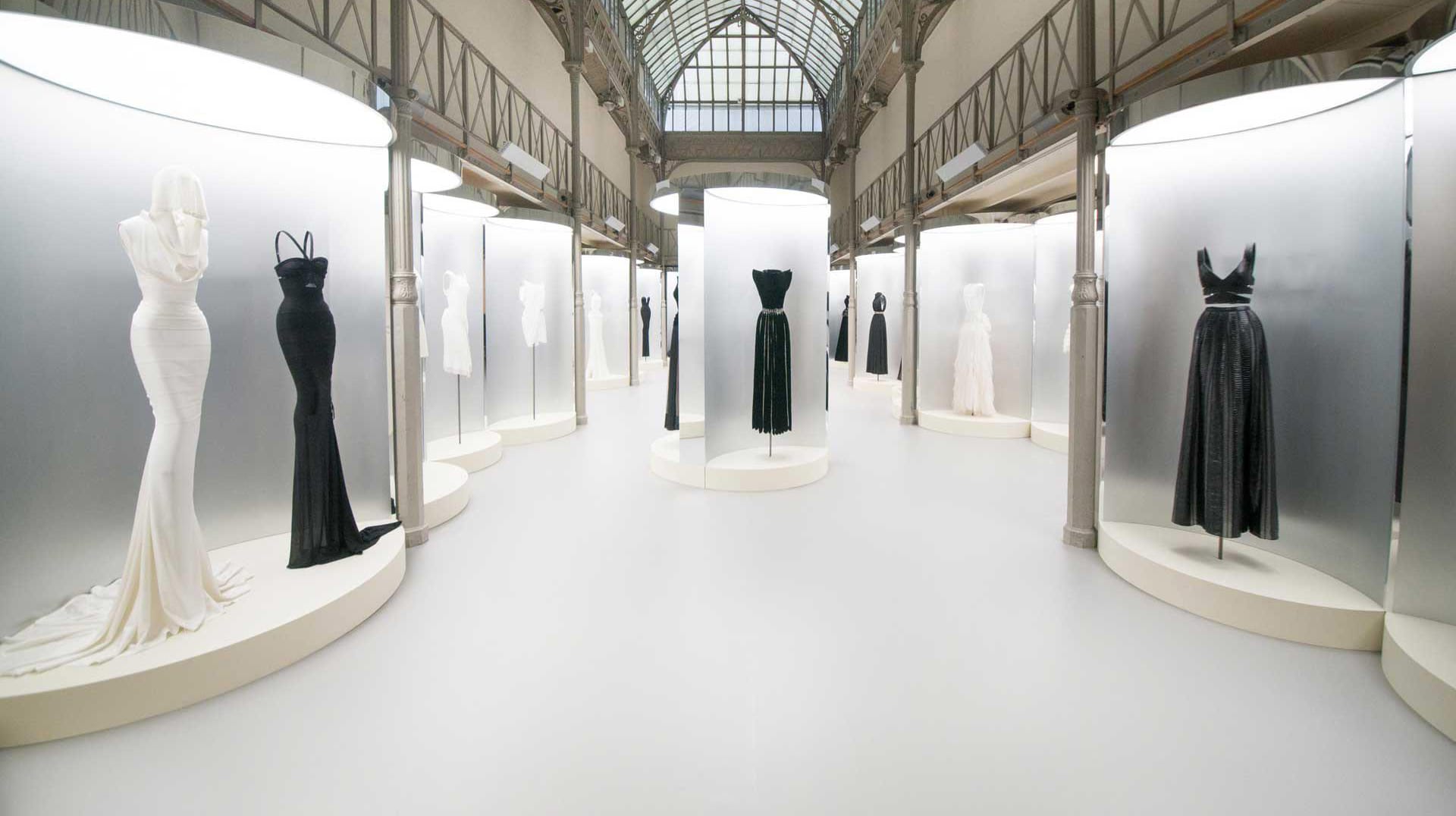 Fashion exhibition with black and white gowns displayed on circular platforms in a spacious, well-lit hall with large windows.