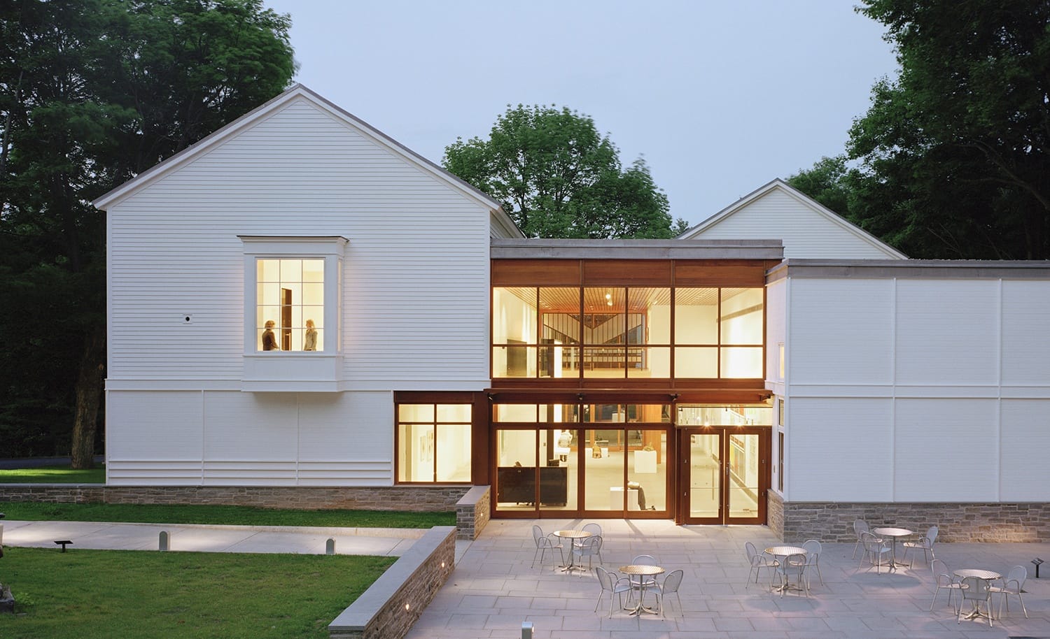 Modern white building with large windows and outdoor patio seating surrounded by trees.