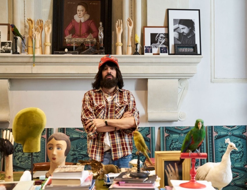 Man standing in eclectic room with artwork, mannequin hands, books, and birds on a table, wearing a plaid shirt and cap.