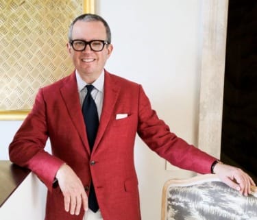 Person in a red blazer with glasses smiling, leaning on a chair in a stylish room.