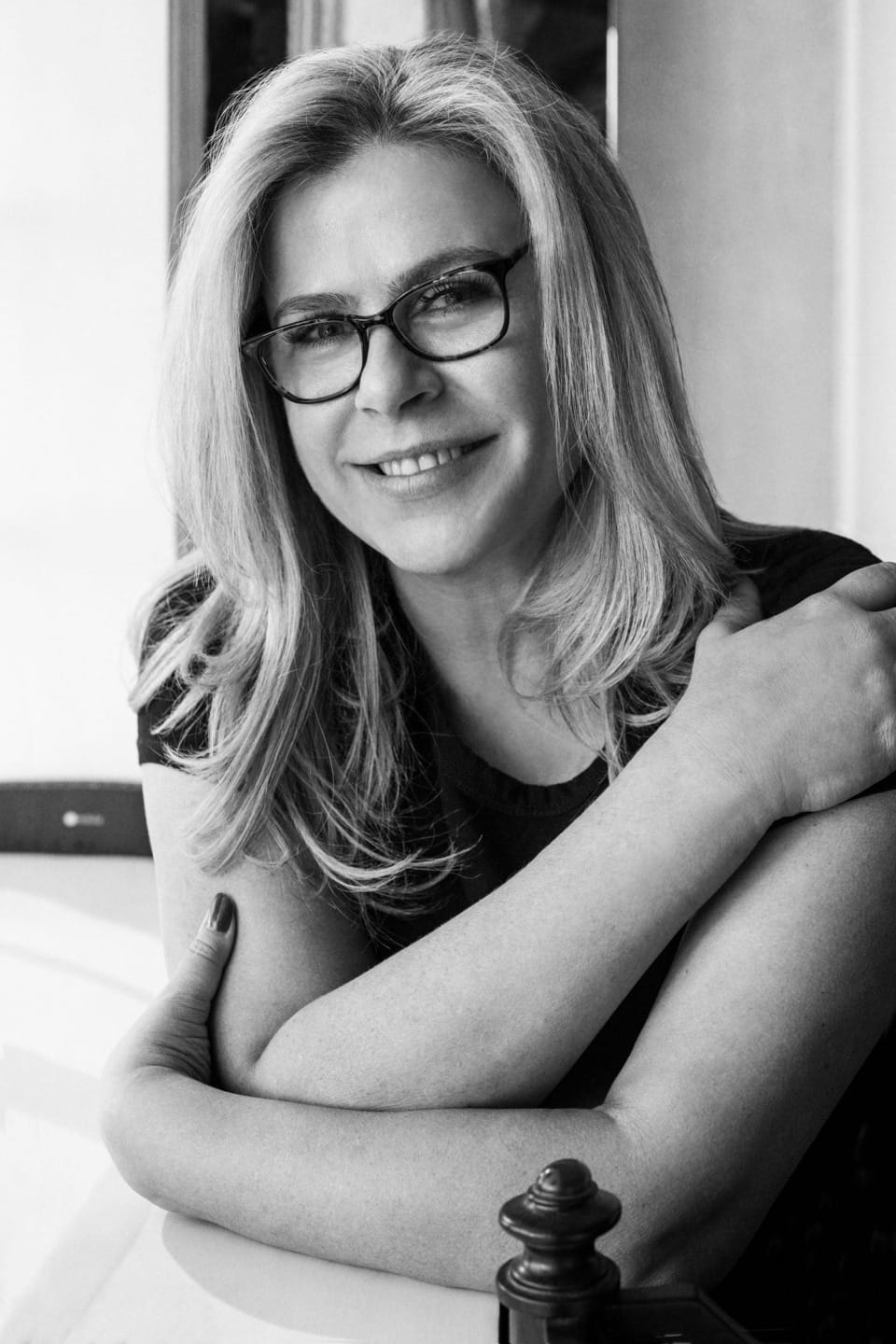 Smiling woman with glasses and long hair sitting at a table, resting her arms, in a black and white photo.