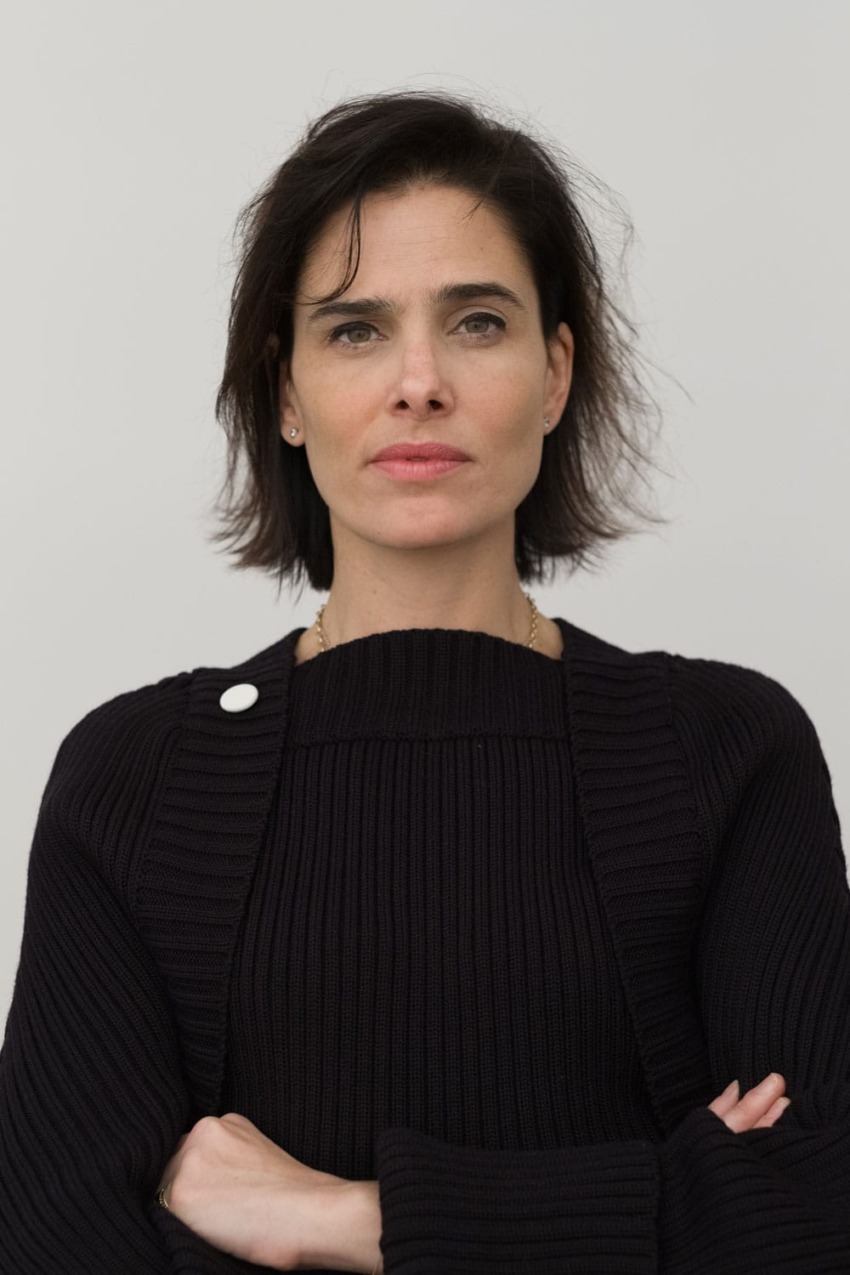 Person with short dark hair wearing a black sweater, arms crossed, neutral expression against a plain background