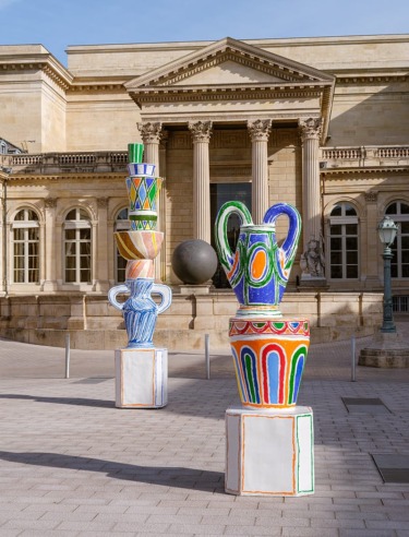 Colorful modern sculptures of oversized vases in front of a historic building with columns and a spherical sculpture.