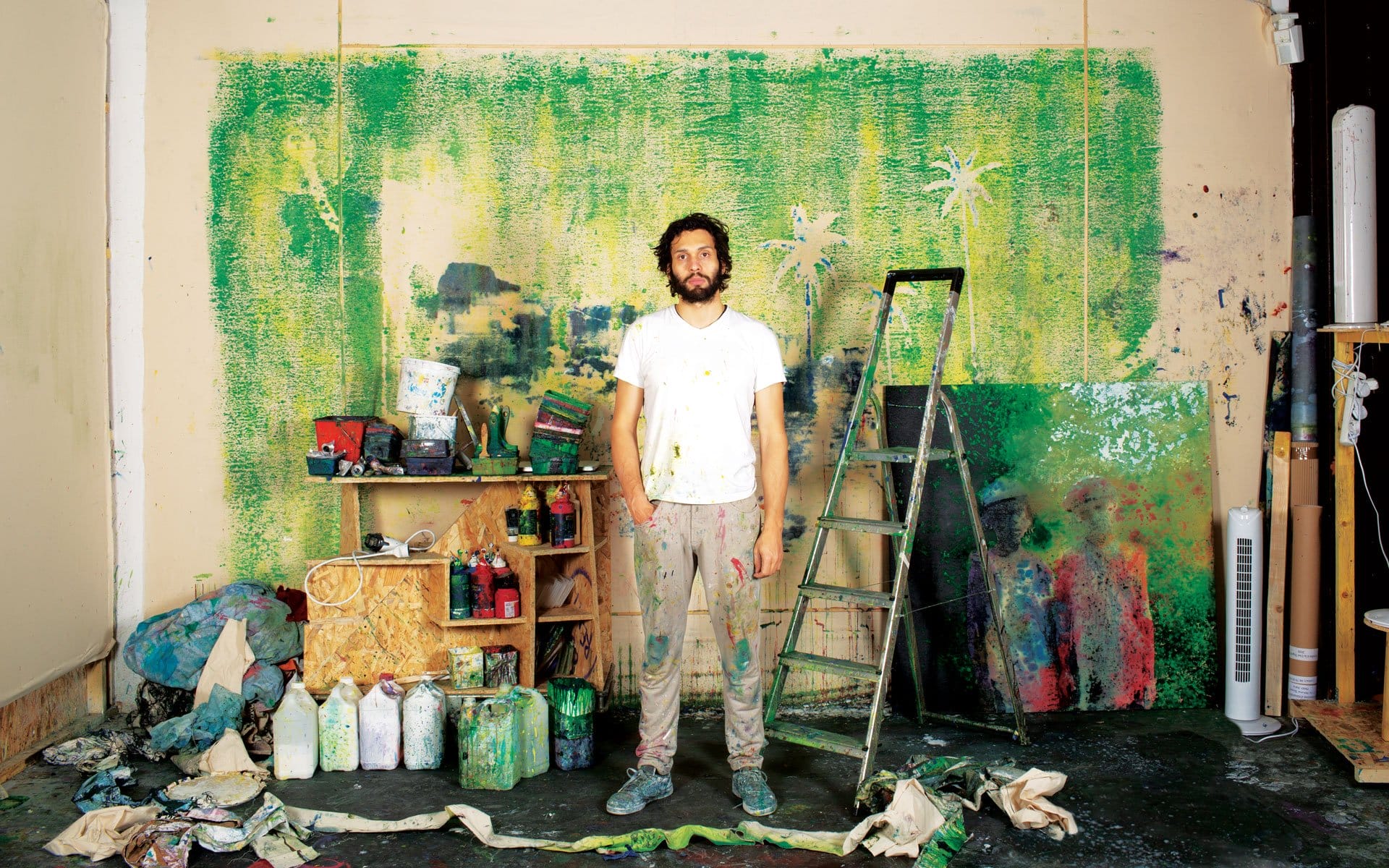 Artist in a paint-splattered studio stands in front of a colorful work-in-progress mural with various tools scattered around.