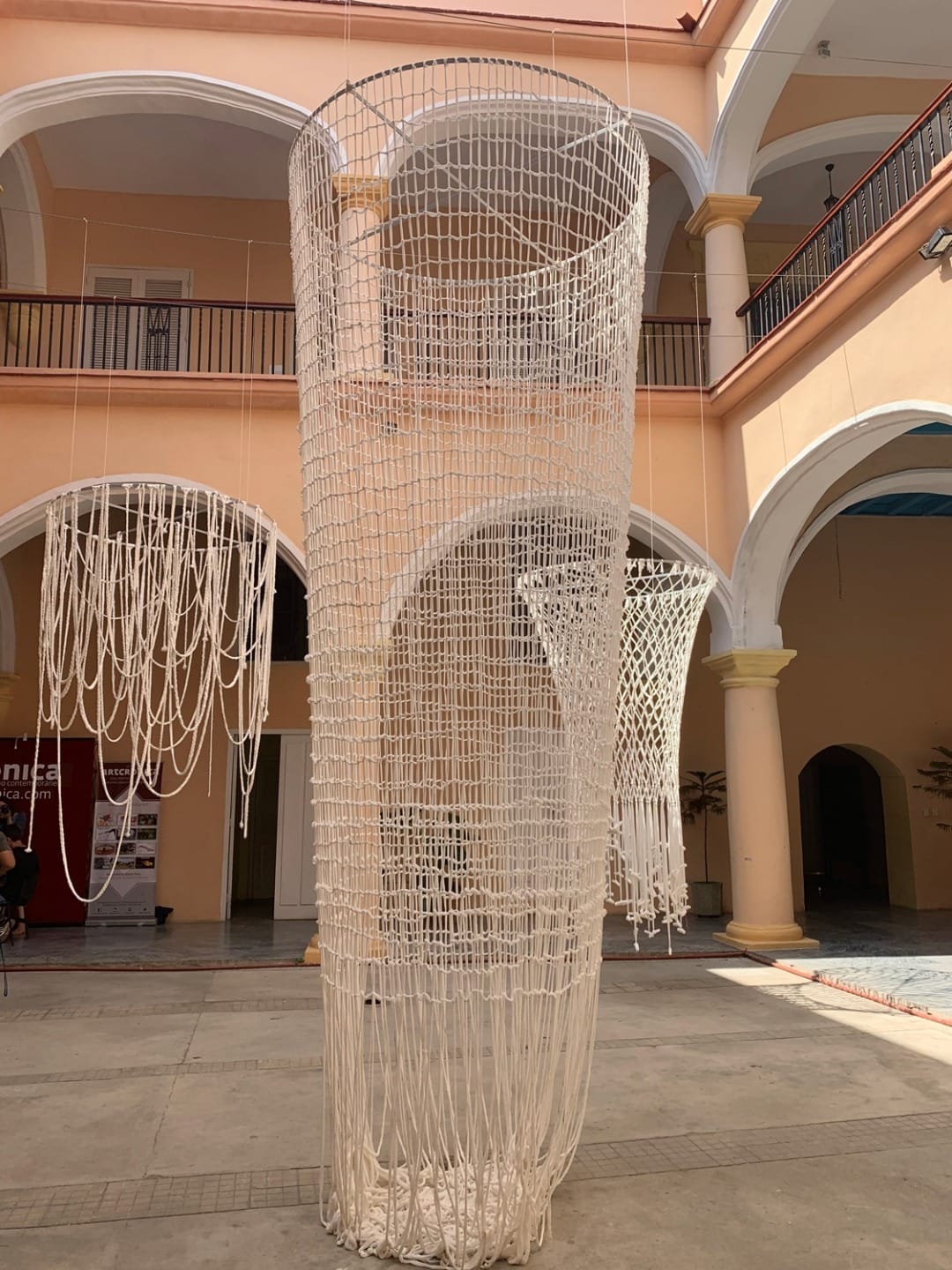 Large white woven cylindrical art installation in a courtyard with arched architecture and high ceilings.